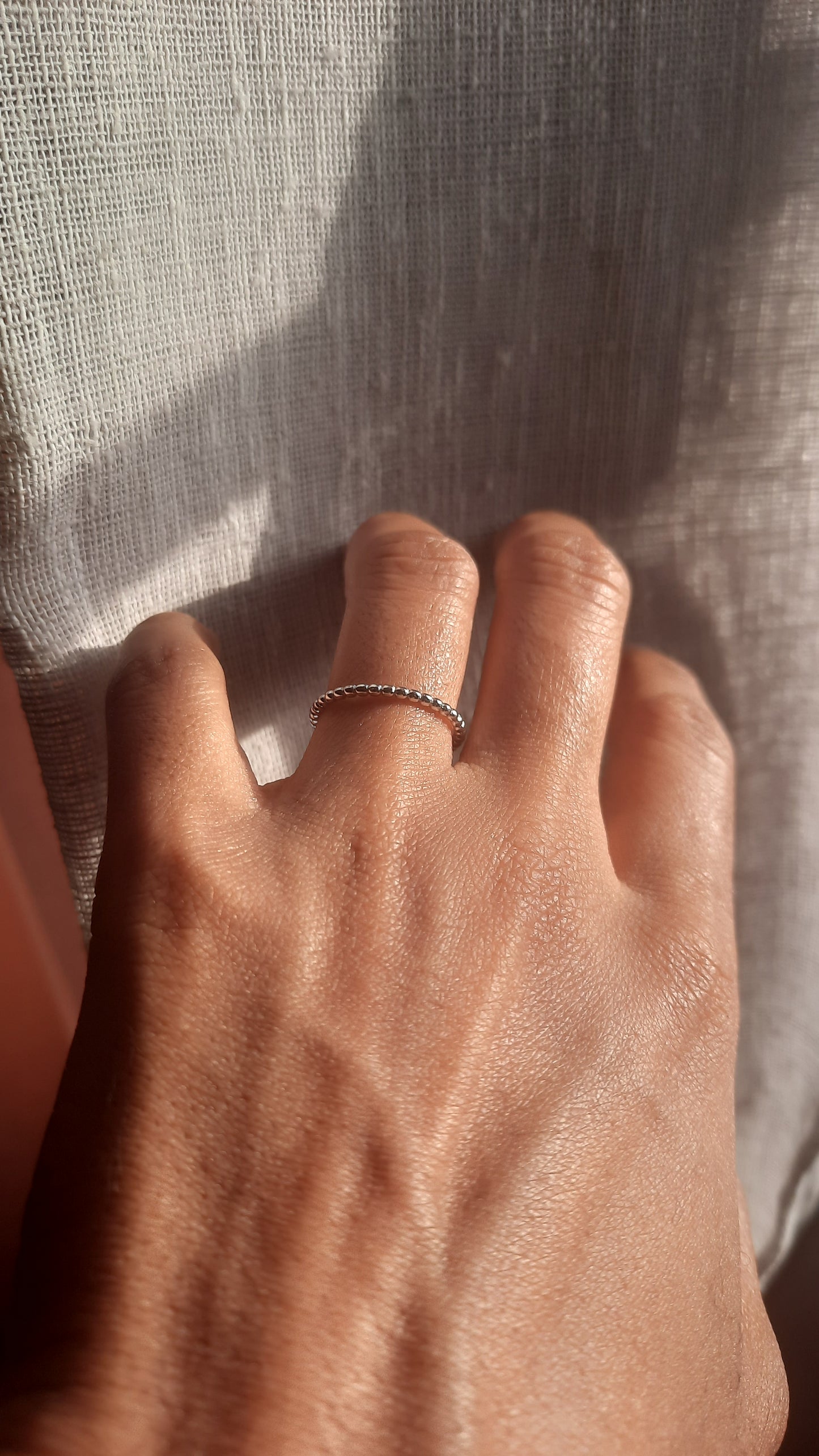 Iliana beaded silver ring on the ring finger of a left hand. The background has sunlight shades over taupe fabric.